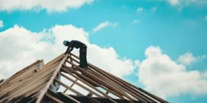 A construction worker balances on a wooden roof against a backdrop of blue sky and clouds